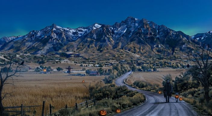 Halloween night in Cache Valley: the Wellsville Mountains under a dark teal sky, a scarecrow in a golden field, bare trees with crows, and glowing jack-o'-lanterns along the dirt road, where a figure in a pale mask and ragged dark cloak walks with two small companions holding jack-o'-lanterns.
