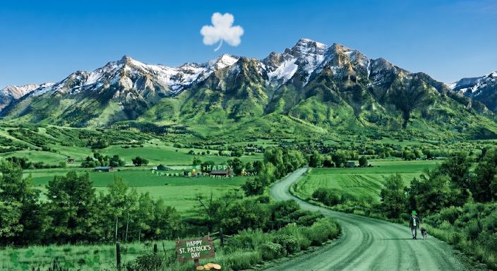 St. Patrick's Day in Cache Valley: a shamrock-shaped cloud over green Wellsville Mountains and fields, a Happy St. Patrick's Day banner by a barn, a wooden Happy St. Patrick's Day sign with gold coins by the fence, and a person in a green top hat walking a dog down the road.